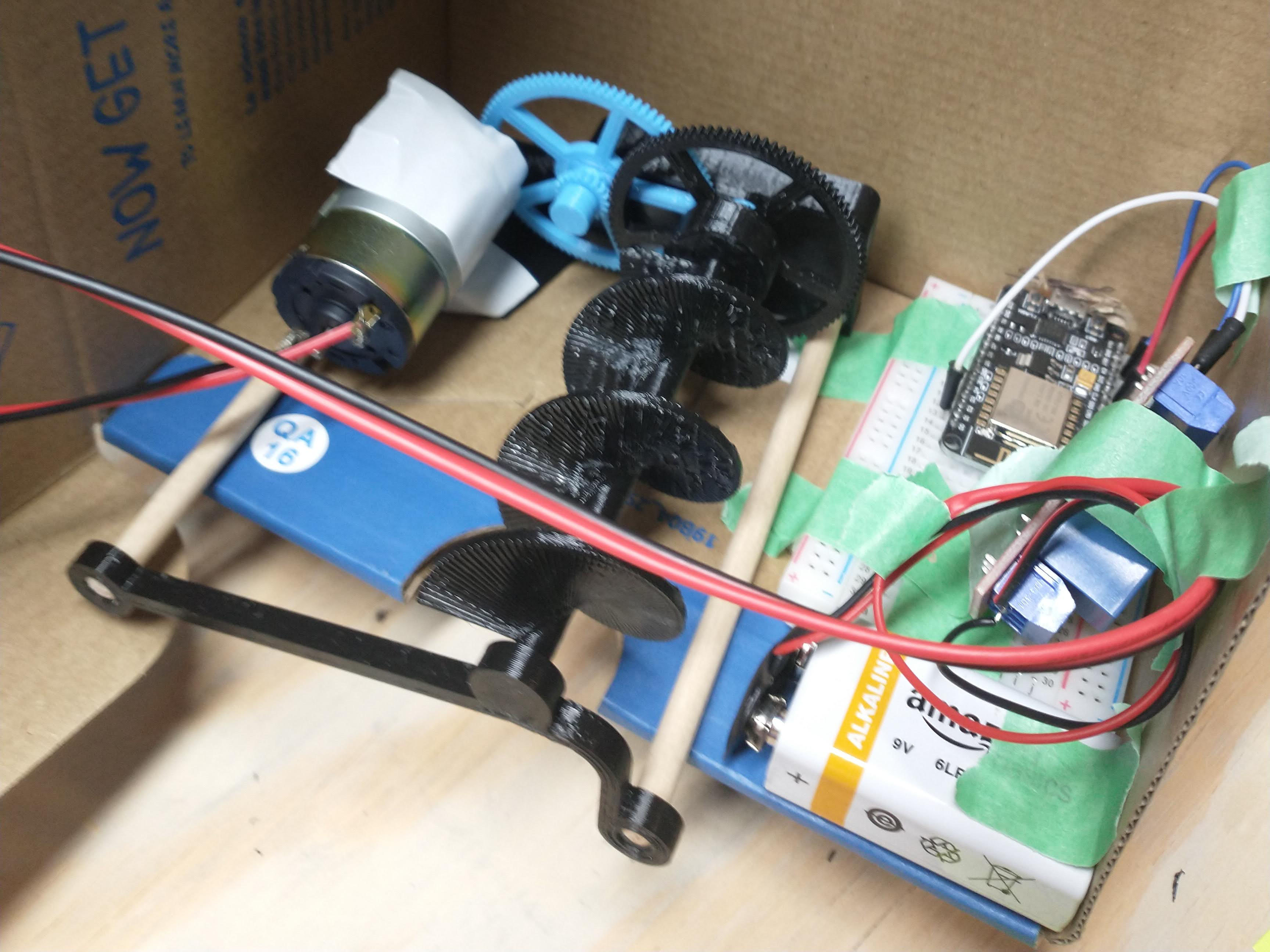 A picture of the electronics driving the cat feeder housed in a cardboard box, featuring an auger connected to a gear connected to a motor, a relay controlling the motor with a 9V battery, and an ESP8266 microcontroller